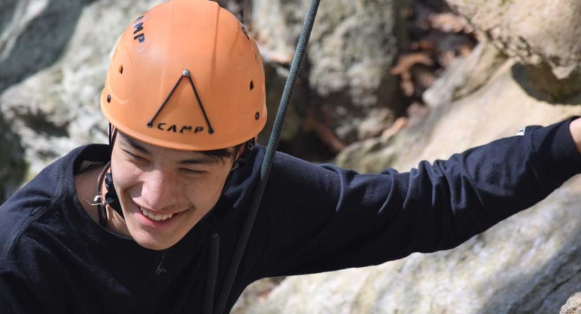 A person wearing a helmet and secured by a rope smiles as they appear to rock climb. 